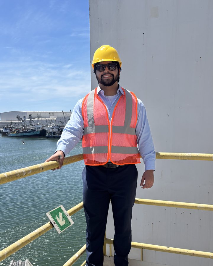 Jesse Regalado on a vessel platform, hard hat and safety vest, fishing fleet behind